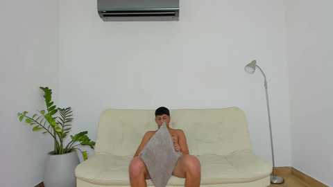 Media: Video of a muscular, shirtless man with short dark hair, wearing only shorts, reading a newspaper on a cream-colored sofa in a minimalist, white-walled room with a potted plant, lamp, and air conditioner.