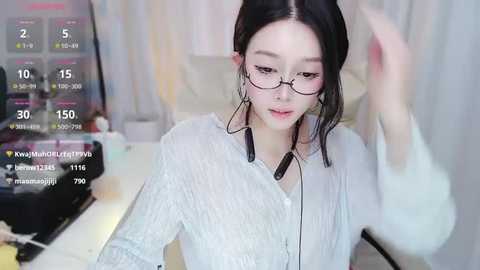 Media: Video of a young Asian woman with long black hair, wearing glasses and a white blouse, adjusting her hair, in a softly lit room with a computer screen displaying weather data.