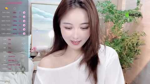 Media: A video of a young Asian woman with fair skin, long brown hair, and a white off-shoulder top, smiling gently, in a well-lit, indoor setting with greenery.