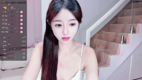 Media: Video of an East Asian woman with long black hair, fair skin, and pink lips, wearing a white camisole. Background features a modern staircase and white walls.