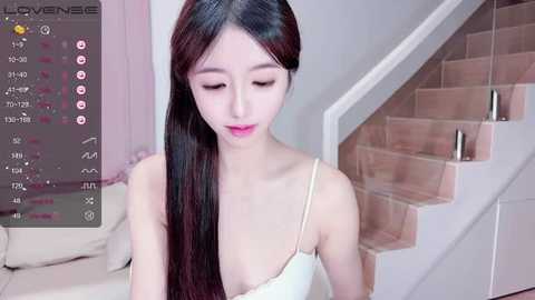 Media: Video of an Asian woman with long, straight black hair, wearing a white spaghetti-strap top, sitting on stairs in a modern, light-colored home.