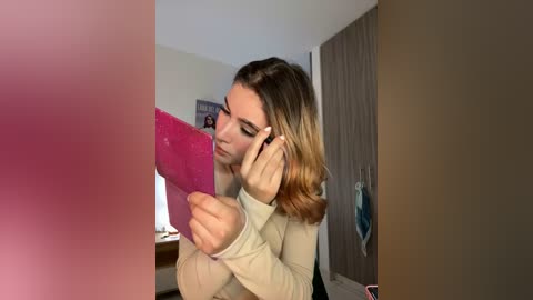 Media: Video of a young woman with shoulder-length blonde hair, wearing a beige top, using a pink phone, in a bedroom with a wooden wardrobe and posters.