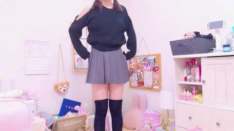 Media: Video of a young woman with fair skin, wearing a black sweater, grey skirt, and black thigh-high socks, standing in a pastel-colored, cluttered bedroom.