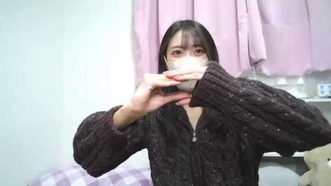 Media: A video of an Asian woman with long black hair, wearing a dark, textured sweater, covering her mouth with a white cloth mask, set in a pastel-colored bedroom with lavender curtains and a white bed.