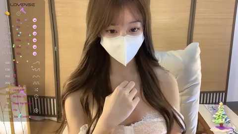 Media: Video of an Asian woman in a hospital room with a white face mask, brown hair, and a lace top, sitting on a bed with a wooden headboard, a calendar, and a toy Christmas tree in the background.