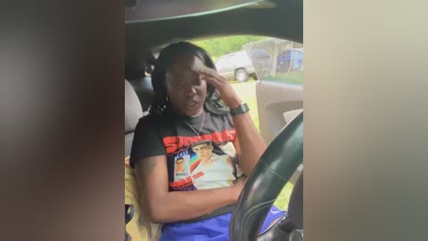 Media: Video of an African American woman in a car, wearing a black t-shirt with colorful graphics, blue shorts, and a black wristwatch. She looks distressed, covering her face with her hand. Outside the window, a blurred parking lot and a car are visible.