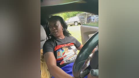 Media: Video of a young woman with short black hair, wearing a dark gray T-shirt with colorful graphic and blue shorts, driving a car, with blurred background of parked vehicles and trees.