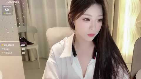 Media: Video of an East Asian woman with long black hair, fair skin, wearing a white blouse, seated indoors, with a blurred background featuring a white chair and soft lighting.