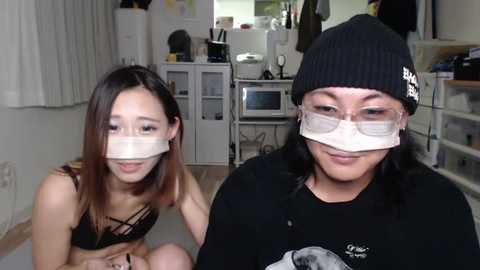 Media: Video of two women wearing face masks in a cluttered, dimly-lit room. One is in a black bra, the other in a black sweater. Background shows a messy kitchen with cabinets and a microwave.