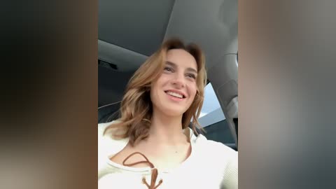 Media: Video of a smiling, light-skinned woman with wavy, shoulder-length brown hair, wearing a white, ribbed knit sweater with a brown bow, seated in a car with gray interior.