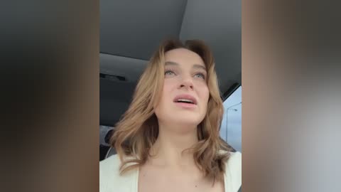 Media: Video of a Caucasian woman with shoulder-length, wavy blonde hair, wearing a white top, standing in a modern room with grey walls and white ceiling.