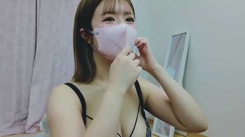 Media: A video of an Asian woman with shoulder-length brown hair, wearing a white face mask, adjusting it with both hands. She has a black bra strap visible. Background includes a lavender curtain and a mirror.