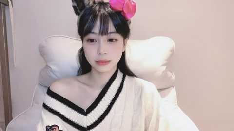 Media: A video of a young East Asian woman with straight black hair, styled with pink macarons, wearing a white off-shoulder sweater with black trim. She sits on a white cushion against a plain beige wall.