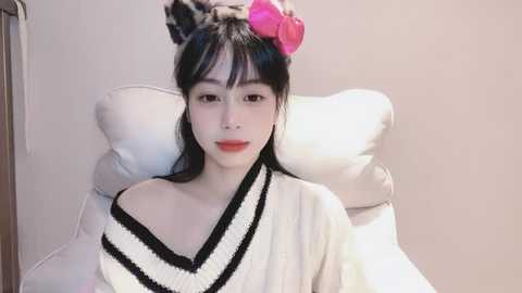 Media: Video of an Asian woman with fair skin, long black hair, and bangs, wearing a white off-shoulder sweater with black trim. She sits on a white pillow against a plain beige wall, with a pink hair clip on her head.