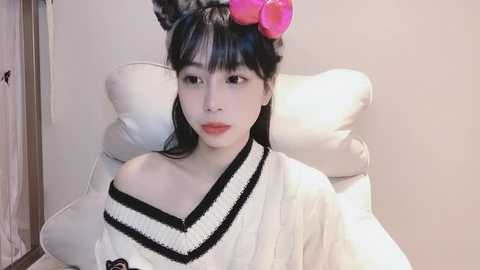 Media: Video of an Asian woman with long black hair, wearing a white kimono with black trim, seated on a white cushion, with a pink fruit on her head, in a simple, indoor setting.