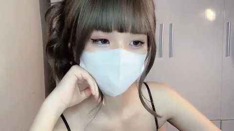 Media: Video of an East Asian woman with long, straight brown hair and bangs, wearing a white face mask and a black spaghetti strap top. She gazes downward, hand near her mouth. Background shows a white wardrobe with vertical handles.