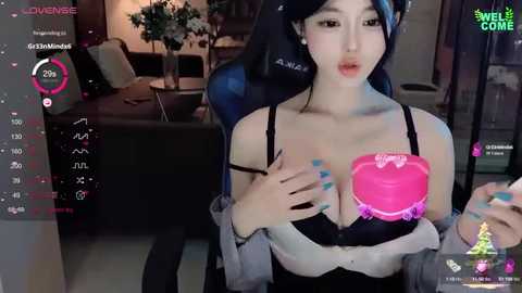 Media: Video of a young East Asian woman with long black hair, fair skin, and large breasts, wearing a black bra and off-shoulder top, holding a pink heart-shaped cake in a gaming livestream setting.