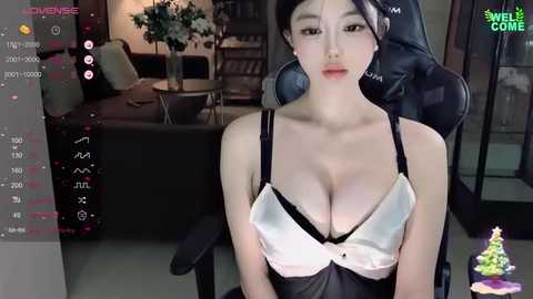 Media: A video of an East Asian woman with fair skin, large breasts, and dark hair, wearing a black and white lingerie set, sitting on a black gaming chair in a dimly lit, modern living room with a TV in the background.