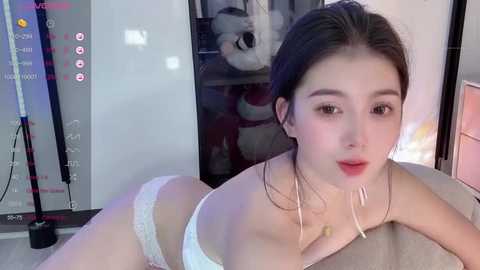 Media: Video of an Asian woman with fair skin and dark hair, wearing white lingerie, leaning forward on a bed in a dimly lit room.