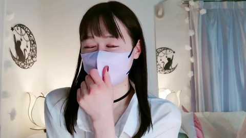 Media: Video of a young Asian woman with long black hair, wearing a white shirt, black choker, and face mask, covering mouth with fingers, in a brightly lit bedroom with white walls, round black decor, and a mirror.