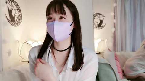 Media: Video of an East Asian woman with long black hair, wearing a light purple face mask and a white blouse, sitting on a bed with pastel-colored pillows and a white bedspread.