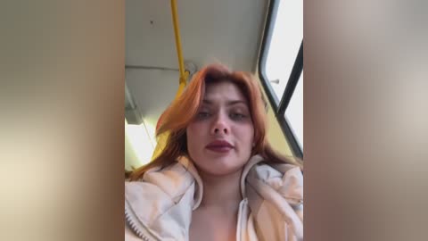 Media: Video of a young woman with long, wavy red hair, wearing a white hoodie, seated in a yellow bus, looking out the window.