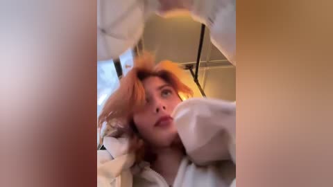 Media: A video of a young woman with red hair, wearing a white robe, looking up in an indoor setting with blurred background.