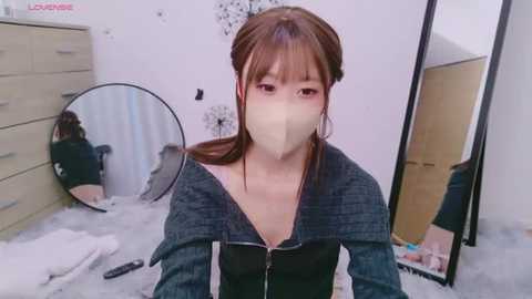 Media: Video of a young Asian woman with fair skin, wearing a mask, black jacket, and long brown hair, sitting on a bed in a messy room with white walls, a mirror, and a dresser.