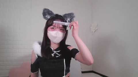 Media: Video of a young woman with pale skin, long black hair, and cat ears, wearing a white face mask, black maid outfit, and holding a hairbrush. She stands in a minimalist white room with a brick wall.