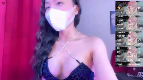 Media: Video of a young Asian woman with long black hair, wearing a white face mask and a black lace bra with crisscross straps. Background features red curtains and a TV screen displaying a video game.