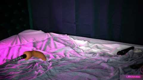 Media: Video of a messy bedroom with a white bed and a person, partially covered by a gray sheet, holding a bottle and remote. Dark curtains in the background, illuminated by pink and purple lights.