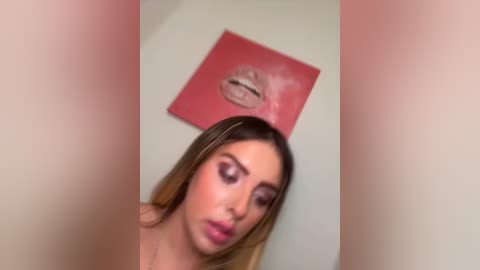 Media: Video of a young woman with long blonde hair and light skin, wearing pink lipstick and eye makeup, against a red wall with a large, circular \"Kiss\" art piece.