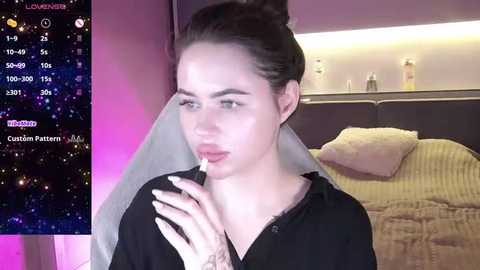 Media: Video of a young woman with light skin and dark hair tied back, wearing a black blouse, looking contemplative while holding a lollipop. Background includes a bed with beige sheets, and a digital interface with a starry sky overlay.