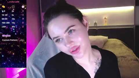 Media: Video of a fair-skinned, dark-haired woman with light makeup, wearing a black top, sitting on a bed with beige pillows, in a dimly lit room, with a custom video pattern in the background.