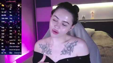 Media: Video of a fair-skinned woman with dark hair in buns, wearing a black off-shoulder top revealing floral tattoos on her chest, sitting in a dimly-lit bedroom with purple lighting and a bed.