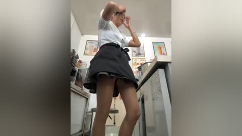 Media: A video captures a woman in a white blouse and black skirt, bending over in a modern kitchen. She has glasses and is holding her head, with kitchen appliances and framed pictures in the background.