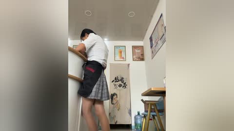 Media: Video of a woman in a white blouse and plaid skirt, cleaning a white wall, in a small, brightly lit room with framed pictures, a water dispenser, and a table.