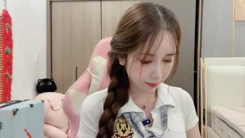 Media: Video of a young Asian woman with long brown hair in a braid, wearing a white polo shirt with a school tie, in a modern, minimalistic bedroom with wooden furniture and a plush toy.