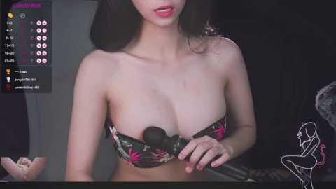 Media: Video of an East Asian woman with long black hair, wearing a strapless floral bra, applying makeup with a black brush. Background includes a dark room with a digital display and a minimalist illustration.