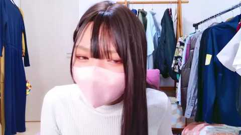 Media: Video of an Asian woman with long dark hair, wearing a white turtleneck and pink face mask, standing in a cluttered closet filled with colorful clothes and a pink purse.