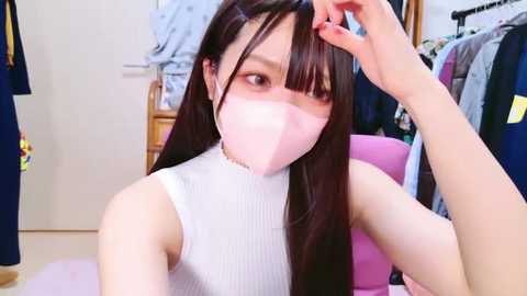 Media: Video of an Asian woman with long black hair and fair skin, wearing a white ribbed tank top and pink face mask, adjusting her hair in a cluttered, brightly lit room.