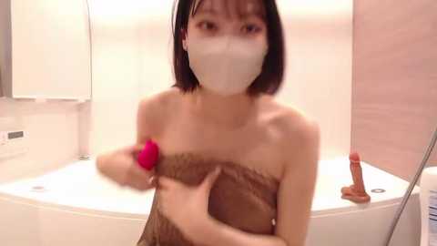 Media: Video of a slender East Asian woman with short black hair, wearing a white face mask and a brown towel covering her breasts. She's holding a pink vibrator and a dildo, standing in a white bathtub with beige walls.