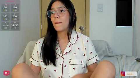 Media: Video of a young woman with long black hair, wearing glasses, and a white pajama top with red flowers, sitting on a bed with white sheets, in a light-colored room.