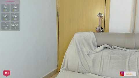 Media: A video of a minimalist, modern bedroom featuring a light gray bed with a beige throw blanket, a wooden door, and a digital clock displaying time and temperature on a light blue wall.