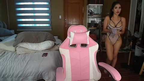 Media: Video of a woman in a revealing, black-and-pink lingerie outfit, standing in a messy bedroom with a pink gaming chair.