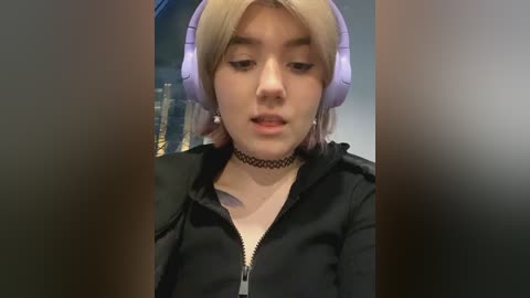 Media: Video of a young woman with fair skin, blonde hair, and large pastel pink headphones. She wears a black hoodie and a black choker necklace, looking down thoughtfully. Background is blurred.