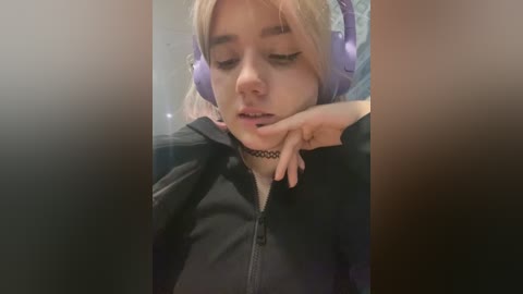 Media: Video of a young woman with blonde hair, wearing purple headphones and a black hoodie, lying with her head on her arm, eyes closed, in a dimly lit room.