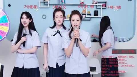 Media: Video of three young East Asian women in white school uniforms, singing into microphones in a brightly lit classroom with a colorful wall calendar and shelves.