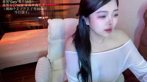 Media: Video of an Asian woman with long black hair, wearing a white off-shoulder top, sitting on a white leather couch in a dimly lit room. Text in Japanese overlays the image.