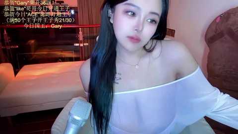 Media: A video of an Asian woman with long black hair, wearing a sheer white off-shoulder top, seated on a bed with a glass of water beside her.